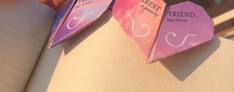 Photo of a two pink-purple origami heart-shaped bookmarks titled 'Dearest Friend, a poem by Rae White', attached to a page of a book, being held by a hand.