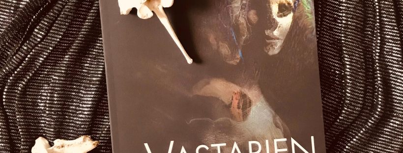 Photo of a dark covered journal with a creepy wooden looking face on the front titled ‘Vastarien A Literary Journal - Vol 3 Issue 2 - Fall 2020’. The journal sits on a black glitter cloth background with two horse spine bones as decoration.