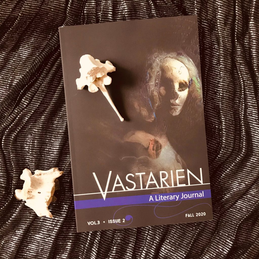 Photo of a dark covered journal with a creepy wooden looking face on the front titled ‘Vastarien A Literary Journal - Vol 3 Issue 2 - Fall 2020’. The journal sits on a black glitter cloth background with two horse spine bones as decoration.