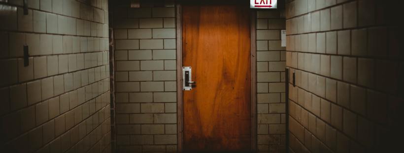 Photo of an orange door in a dimly lit, spooky-looking tiled room, a glowing exit sign in the right hand corner.