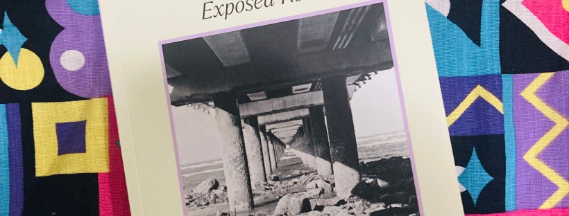 A photograph of the cover of 'Jacaranda: The Emerging Writer's Journal,' Volume 11.1, Winter 2024 edition titled 'Exposed Roots.' The cover features a black-and-white photo of an underpass with columns extending into the distance. The journal is placed on a colourful patterned fabric background.