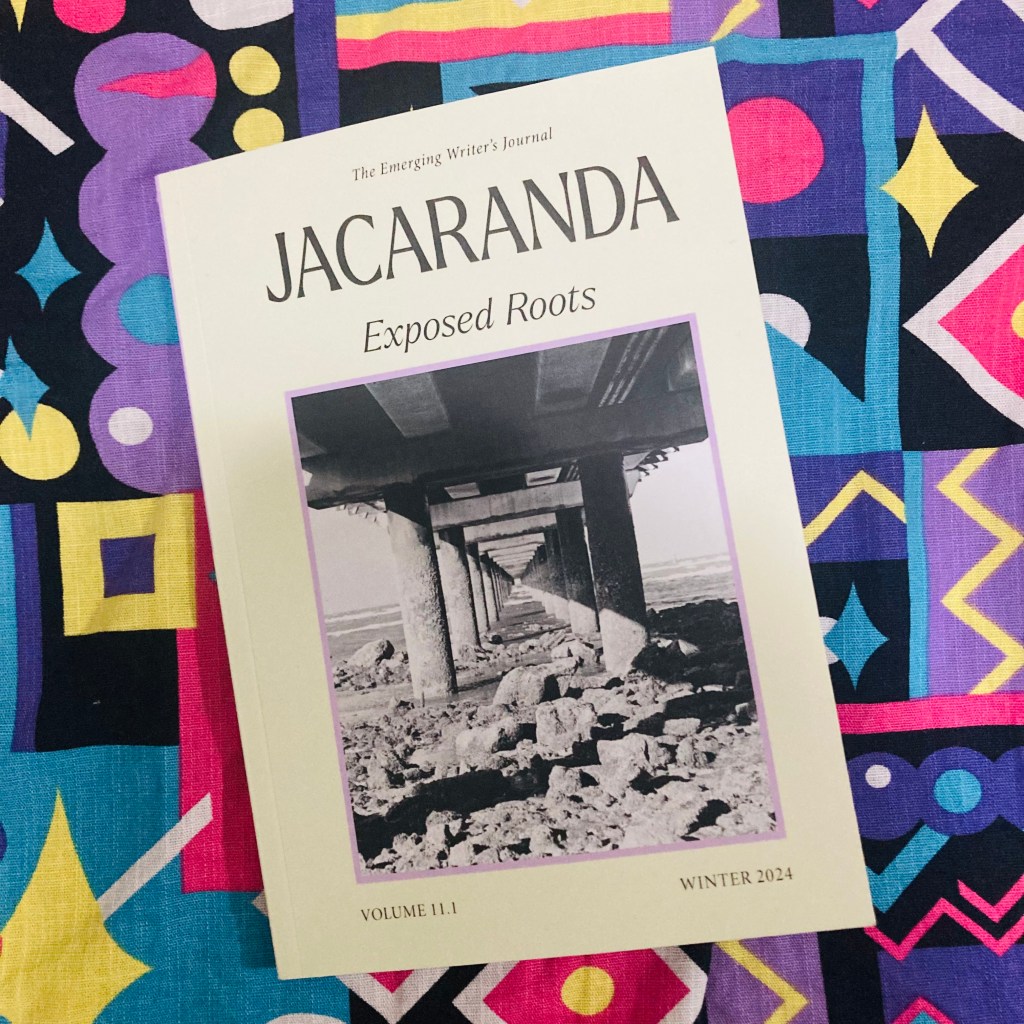 A photograph of the cover of 'Jacaranda: The Emerging Writer's Journal,' Volume 11.1, Winter 2024 edition titled 'Exposed Roots.' The cover features a black-and-white photo of an underpass with columns extending into the distance. The journal is placed on a colourful patterned fabric background.