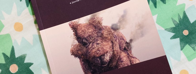 Photo of Rabbit: a journal for nonfiction poetry, Issue 40 titled ‘Extinction,’ placed on a green and cream floral-patterned fabric. The cover of the journal has a dark maroon border, with the central artwork depicting a textured, moss-covered koala with a baby on its back, with smoke rising from its body.