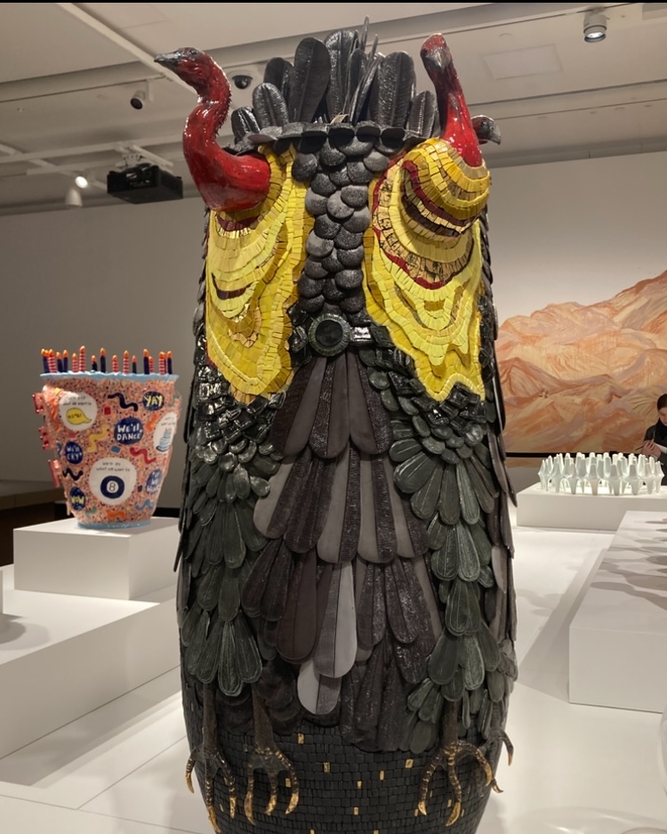 Photo of a sculpture of a multi-headed scrub turkey displayed in an art gallery. The sculpture is covered in black, feather-like textures with intricate mosaic type detailing, and yellow, red and black colouring.